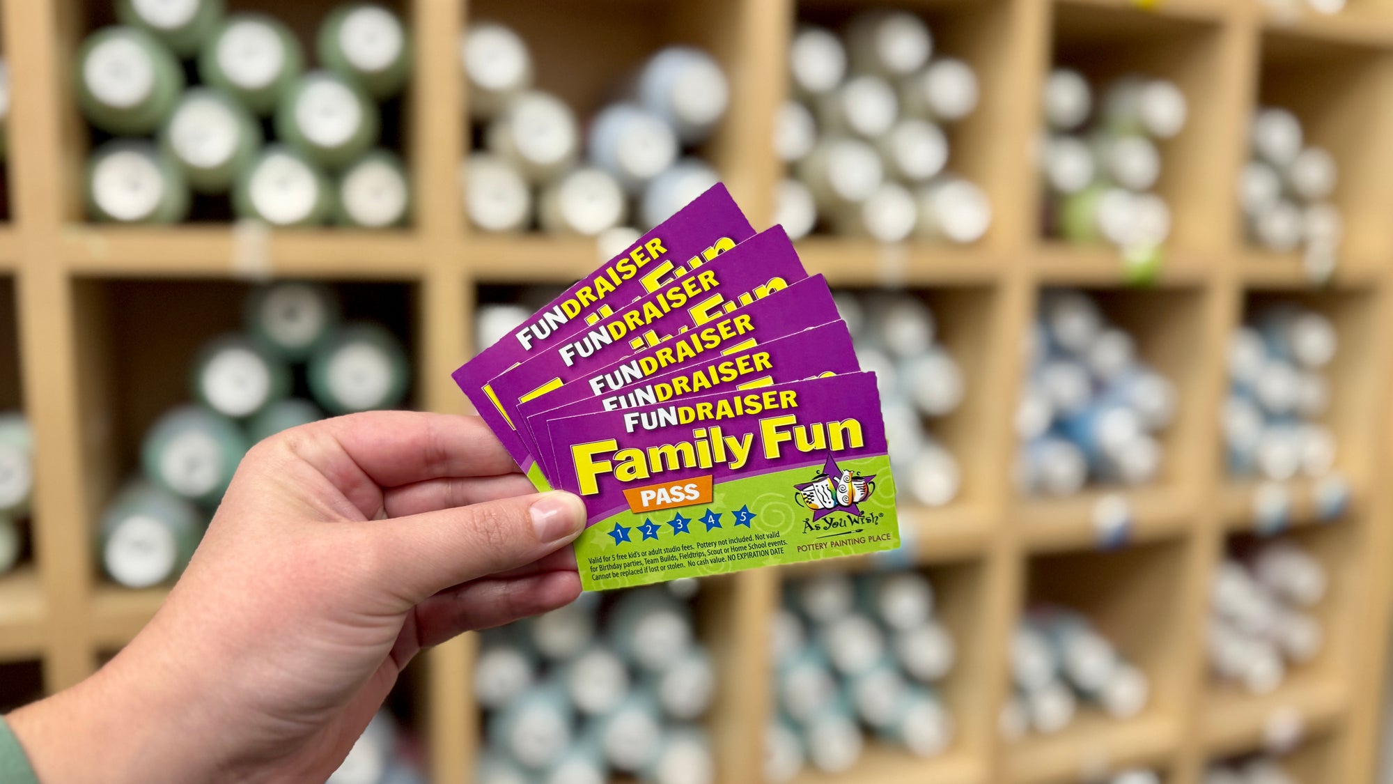 Hand holding Fundraiser Family Fun Passes in front of a paint center with bottles of paint.