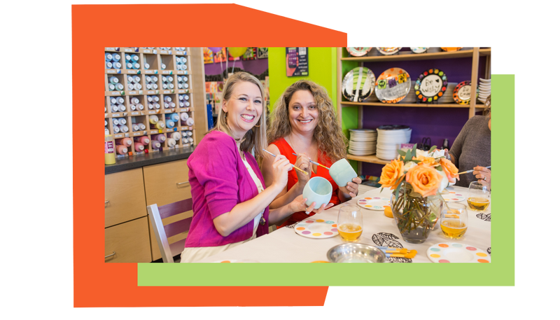 Two women at a table with ceramic items and a colorful background attending a bridal shower at As You Wish Pottery