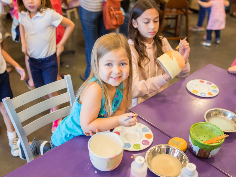 Children at a table with art supplies, including bowls and paintbrushes, in a studio setting.