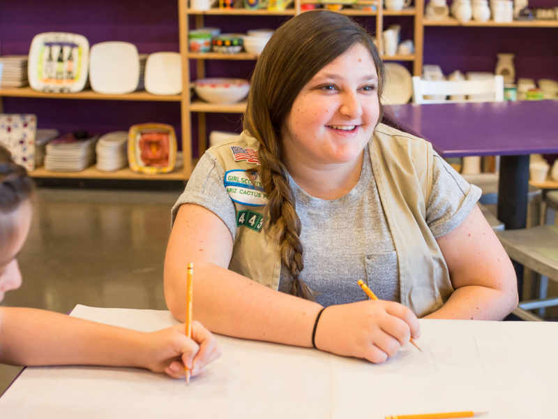 Girl with a Girl Scout uniform sitting at a table with another person, both holding pencils.