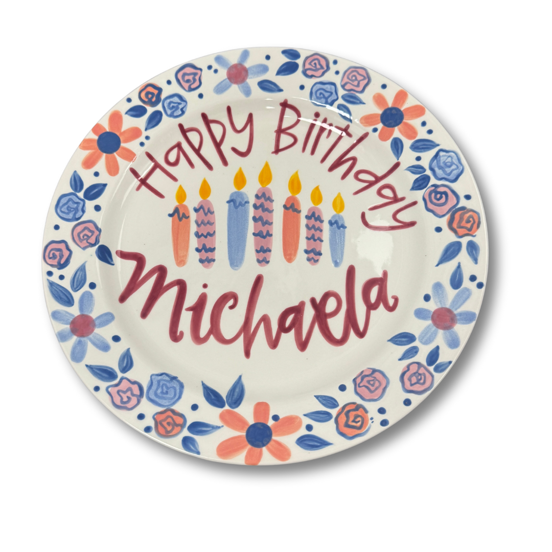 Round ceramic plate with 'Happy Birthday Michaela' text, colorful flowers, and candles on a white background