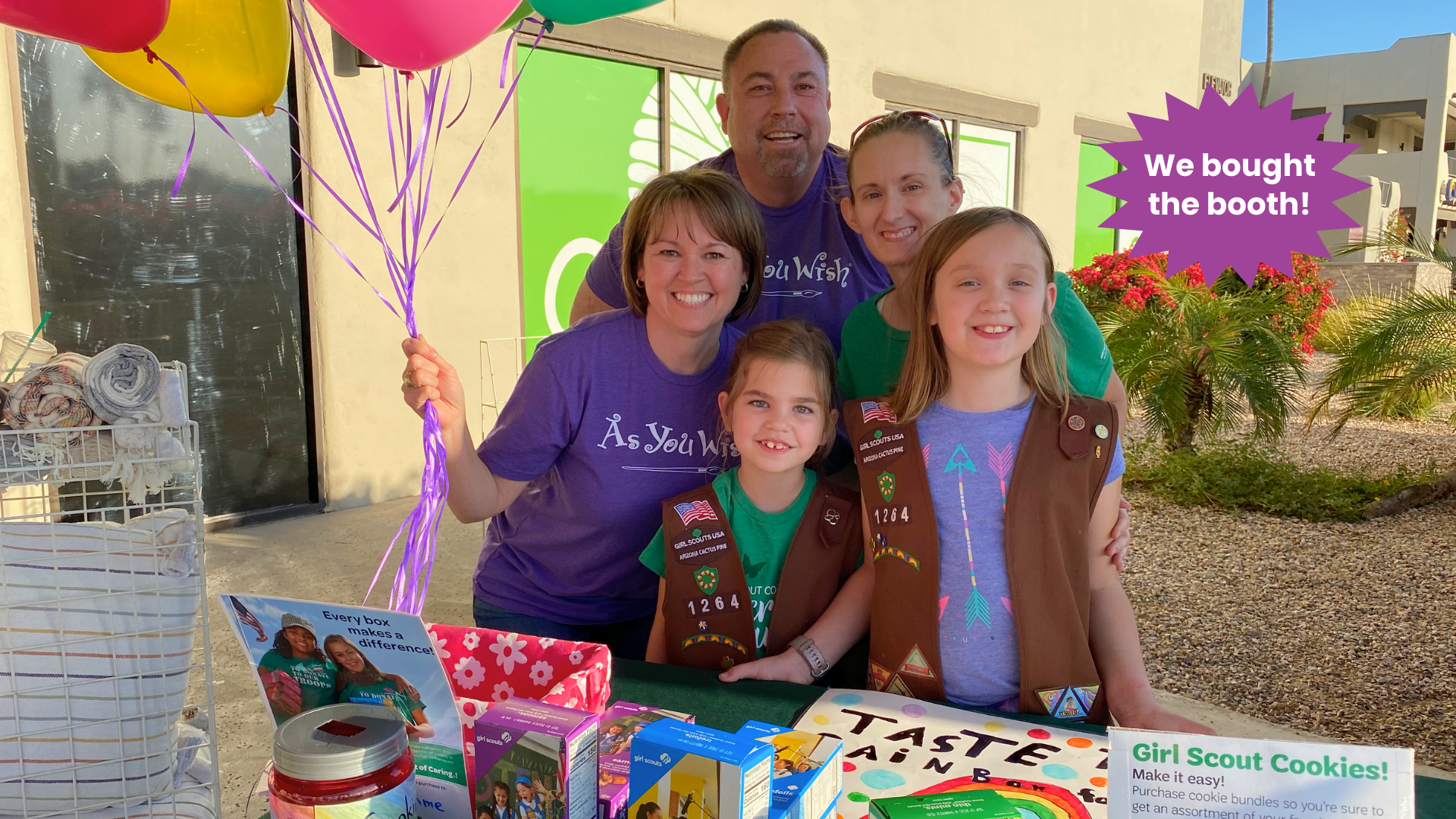 Three adults and two girl scouts with Girl Scout cookies and balloons in front of a booth sign.