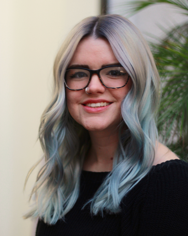 Person with light blue hair and glasses smiling outdoors