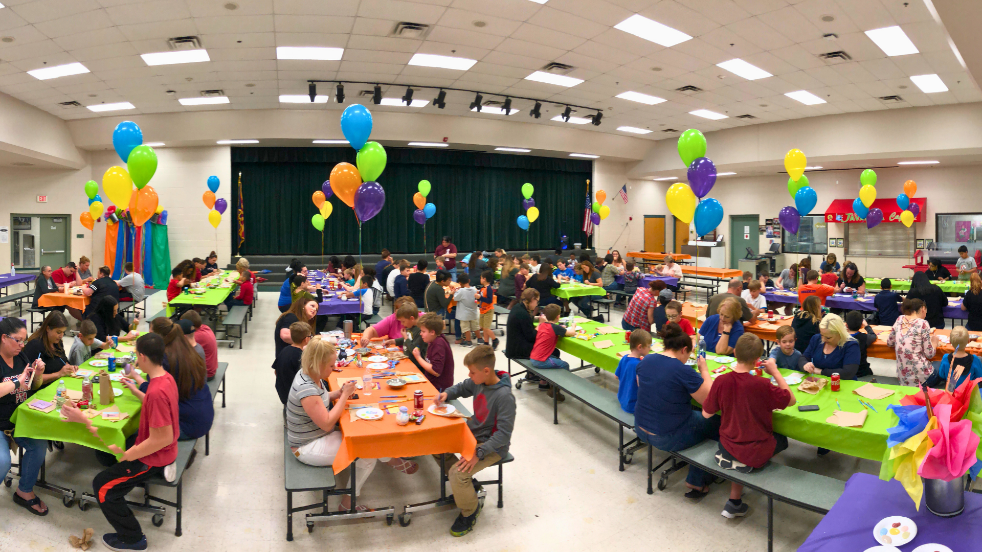 Families in a cafeteria with colorful balloons and tables set up for a pottery painting off-site event.