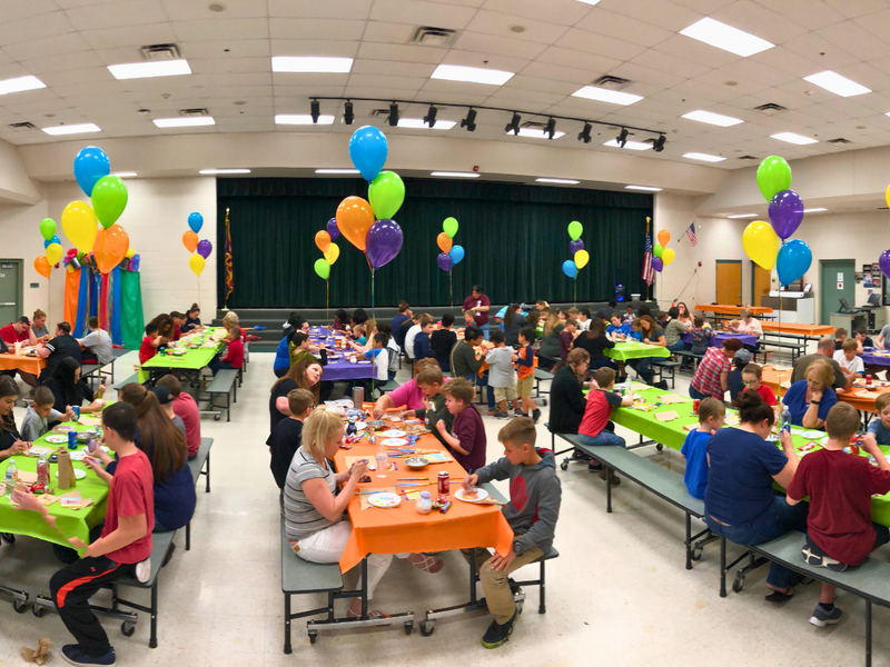 Children sitting at tables painting pottery with colorful tablecloths and balloons in a school cafeteria.