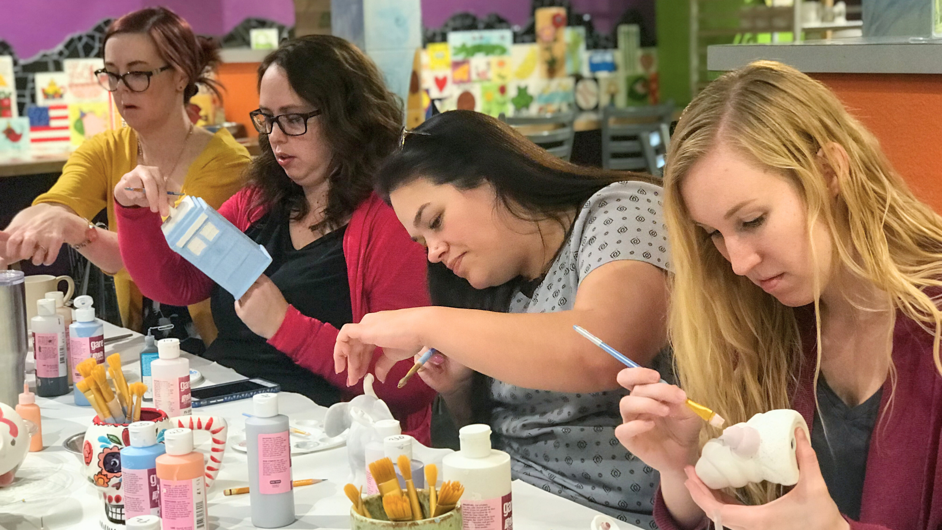Four women engaged in a pottery painting activity at a table with various supplies.