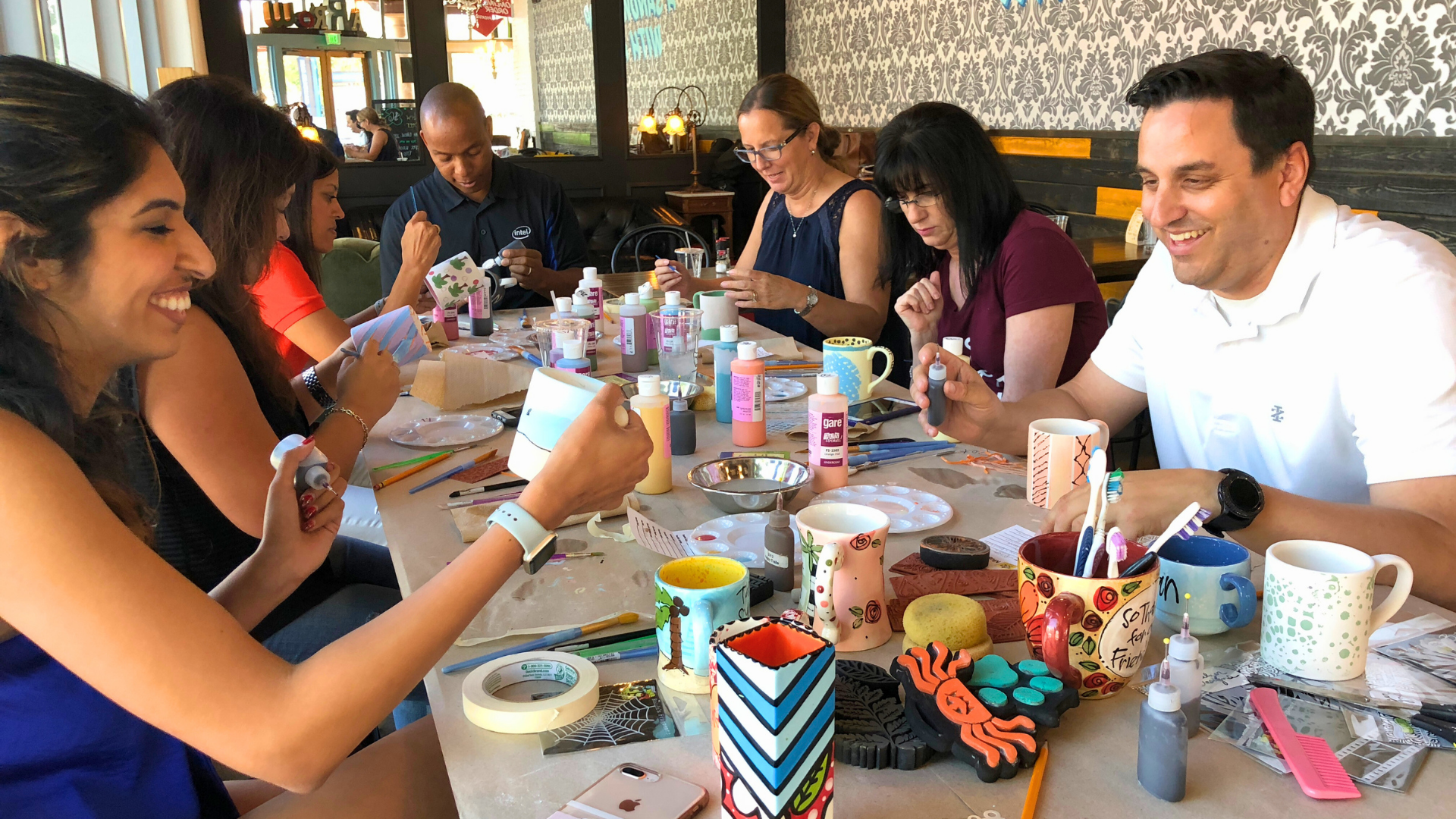 Group of people at a table engaging in a pottery painting team build, surrounded by various craft materials and utensils.