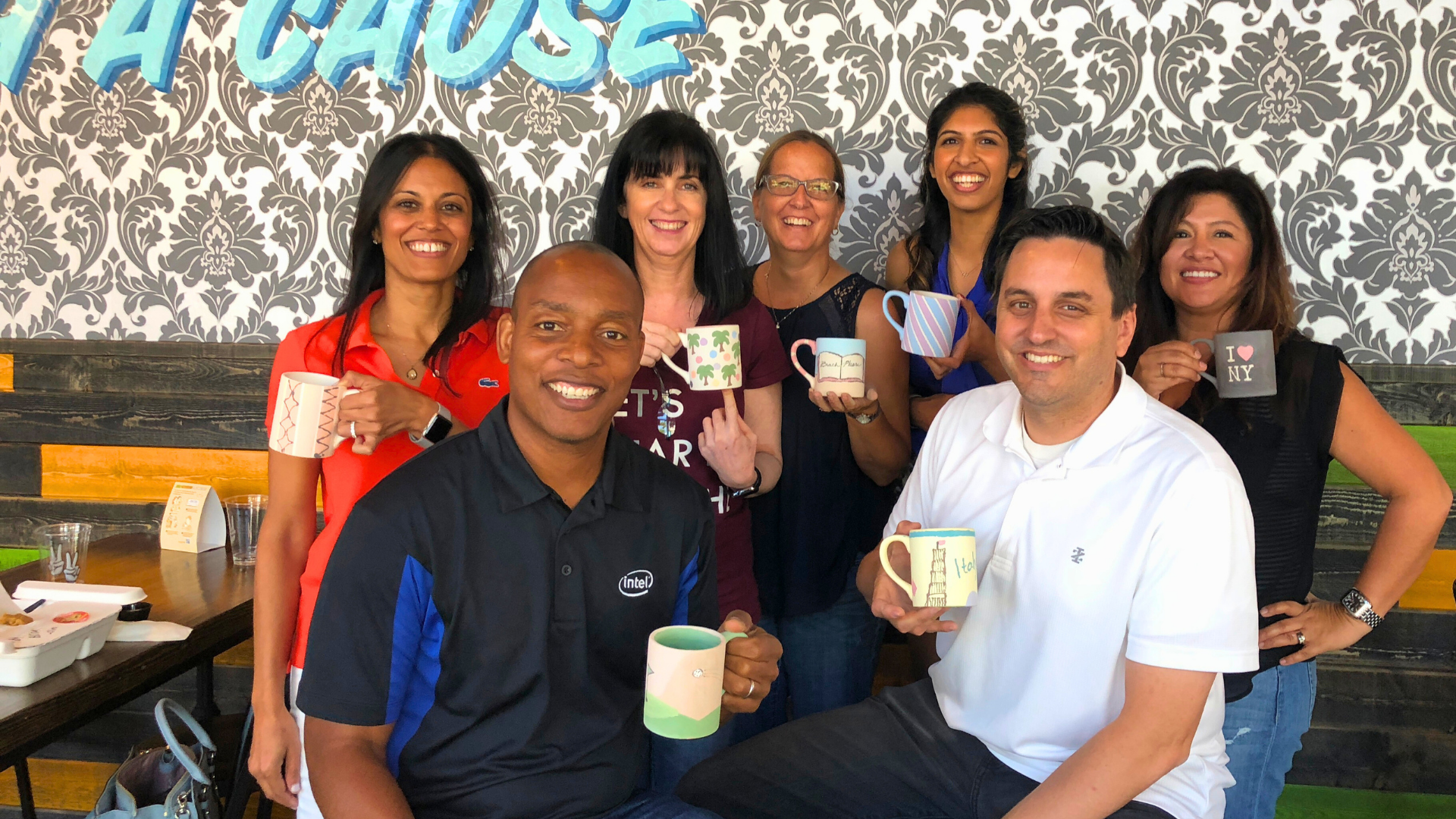 Team of people at an off-site team build holding painted mugs in a casual setting with a decorative wall in the background.